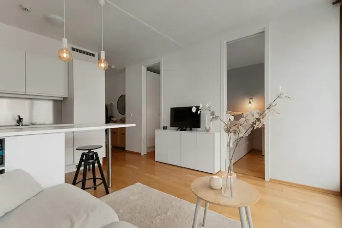 Un salon moderne avec un canapé gris, une table basse en bois, un meuble TV blanc, et une cuisine ouverte avec des placards blancs et des tabourets de bar noirs.