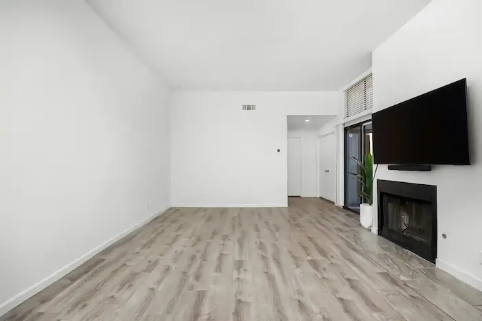 Un salon minimaliste avec un sol en bois clair, des murs blancs, une cheminée encastrée avec une télévision au-dessus, et une porte vitrée menant à un balcon.